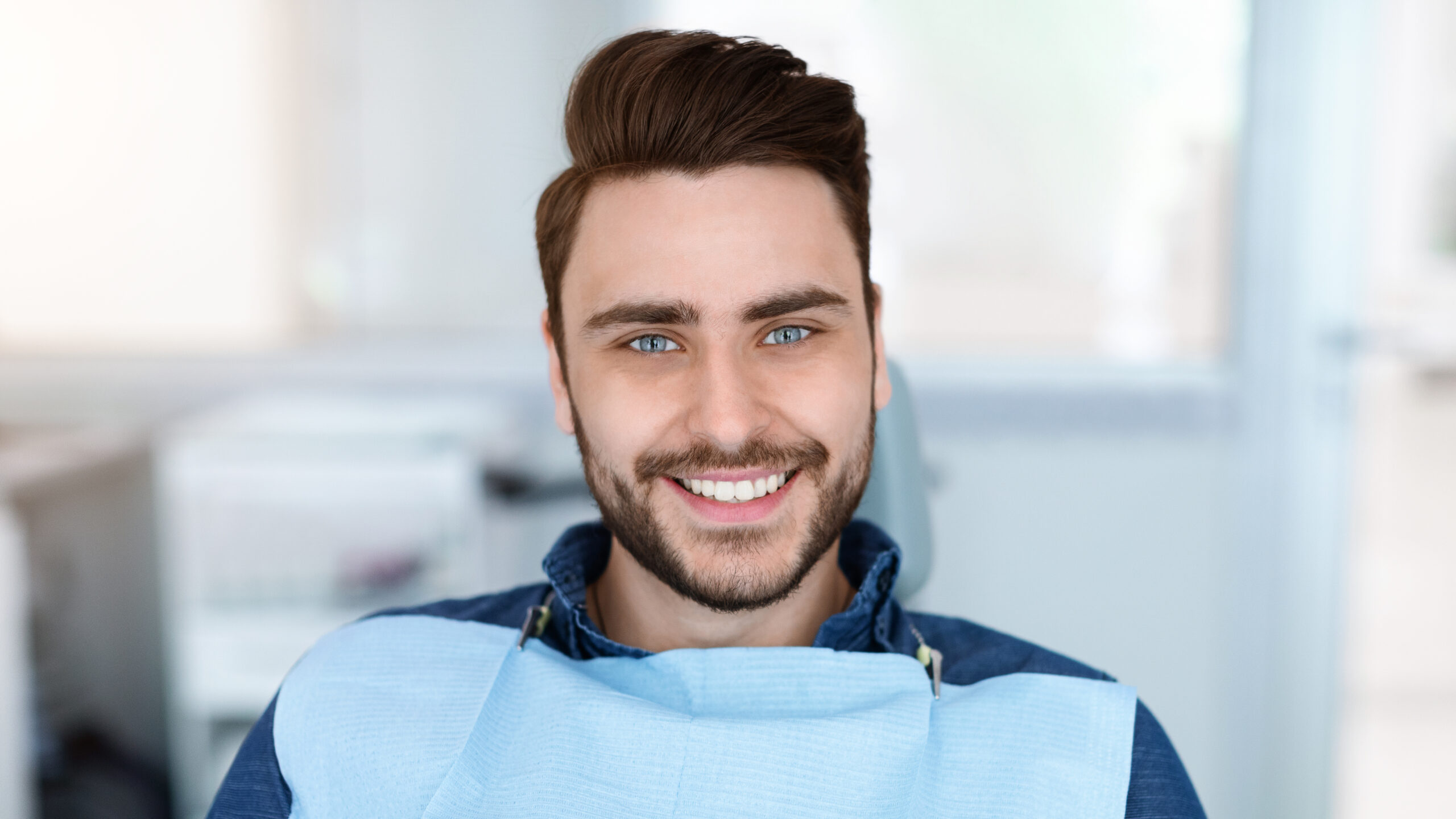 Cheerful bearde man patient sitting in dental chair and smiling at camera, copy space. Positive millennial guy showing his beautiful white smile after treatment at modern dental clinic