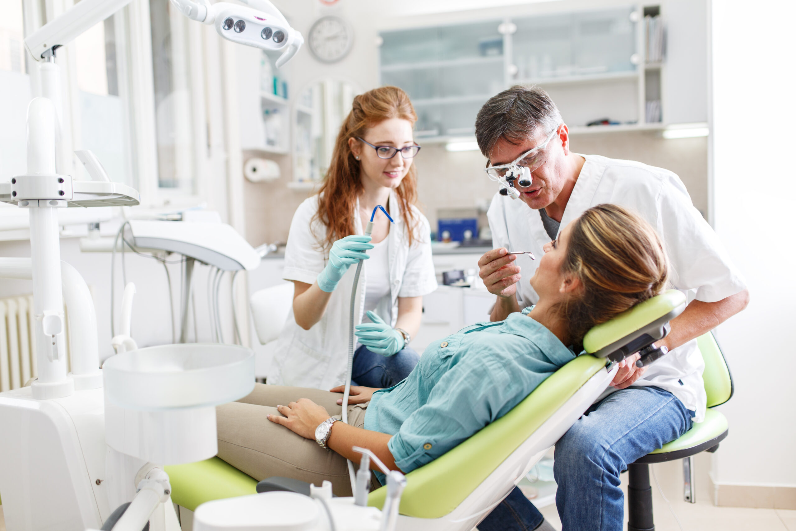 A young woman, seated in the dentist's chair, prepares herself for treatment, her trust in capable hands. Meanwhile, a two dentists discusses her care to ensure a seamless and comfortable experience.